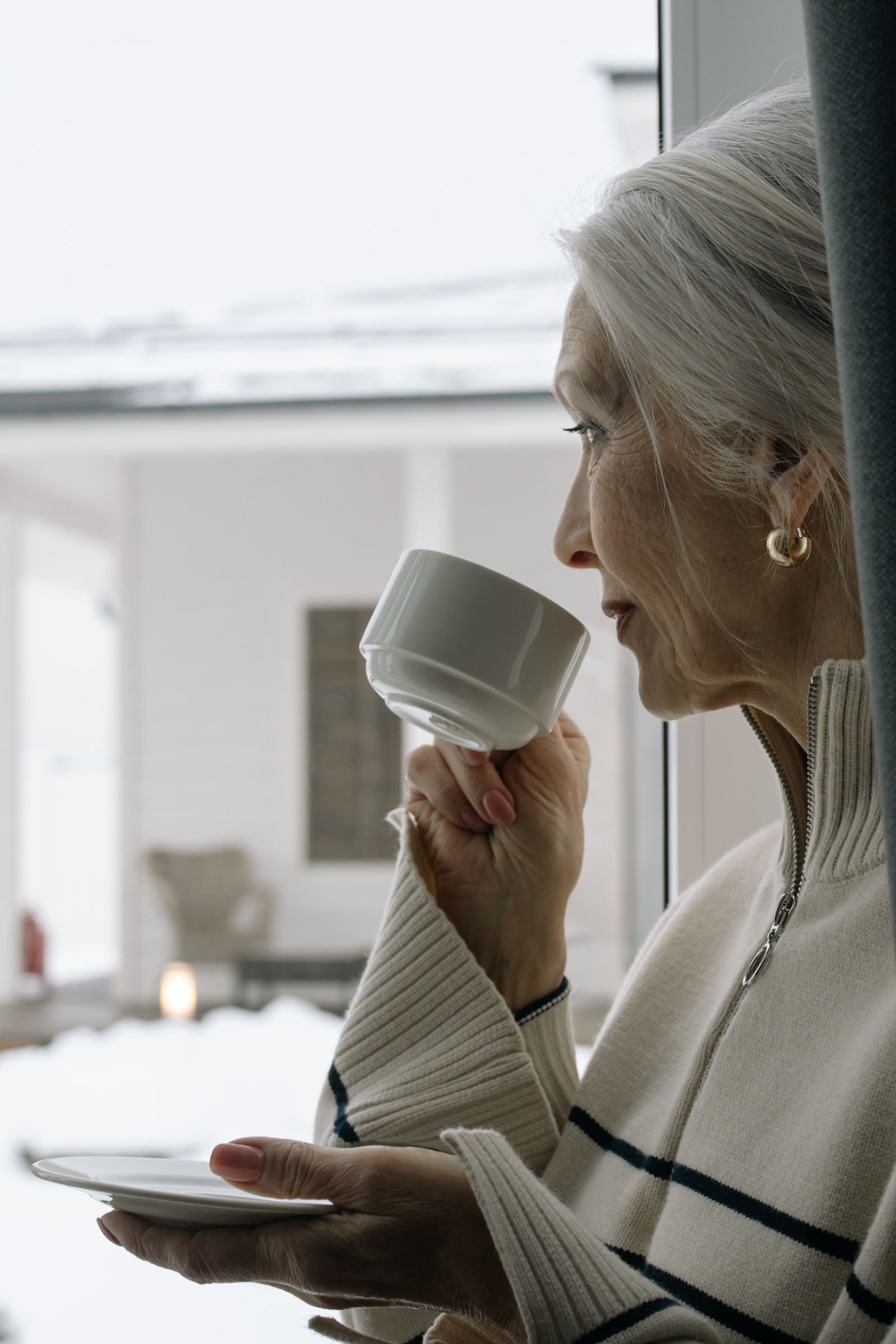 Eldre kvinne drikker kaffe ved vinduet med utsikt mot snødekt tun